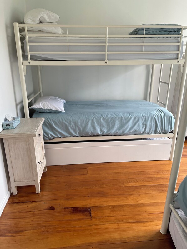 Un lit superposé avec un lit en bas et un lit en haut. Il y a une petite table de chevet avec une boîte de mouchoirs et un tiroir. La pièce a un plancher en bois et des murs blancs.