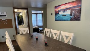 Un comedor con mesa de madera y sillas blancas, un cuadro de una cascada y un tablón de corcho con notas.