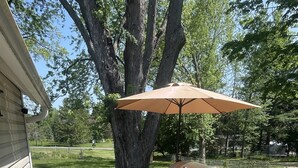 Una terraza con una parrilla y dos sillas de mimbre, rodeada por una barandilla de madera y un árbol grande.
