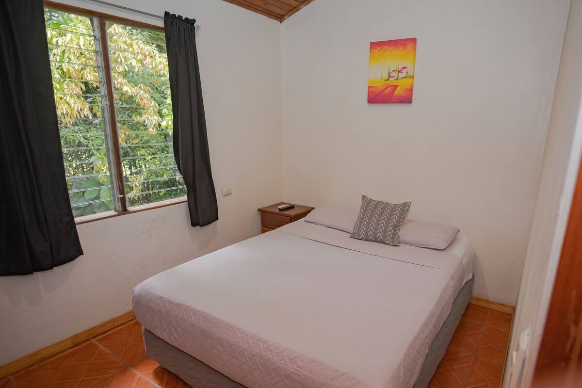 Une chambre à coucher avec un lit, une table de chevet, une fenêtre avec des rideaux et un tableau au mur.