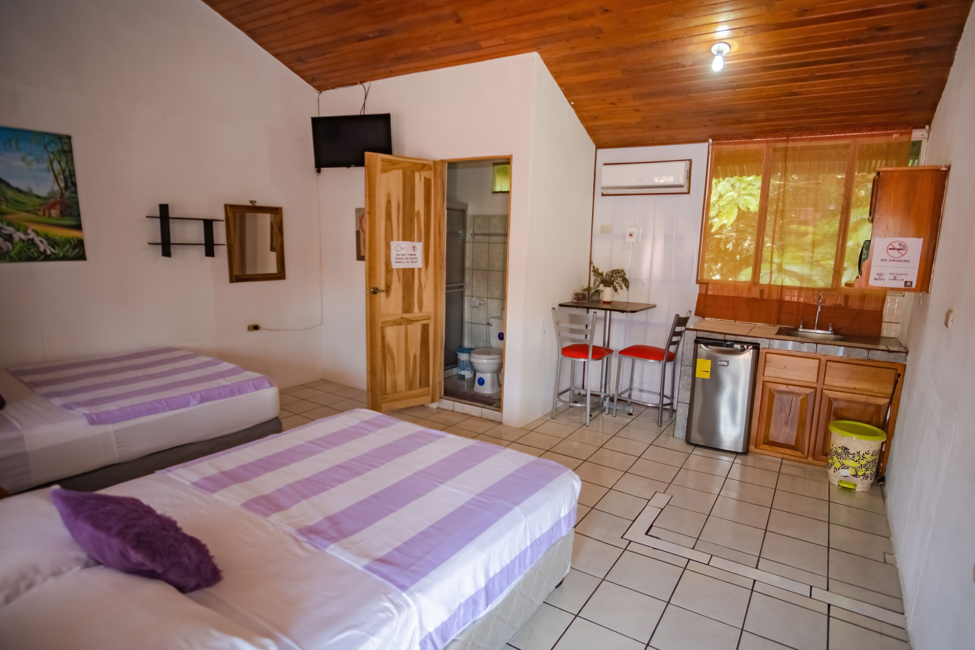 A room with two beds, a wooden ceiling, a kitchen area with a refrigerator, and a bathroom with a shower.