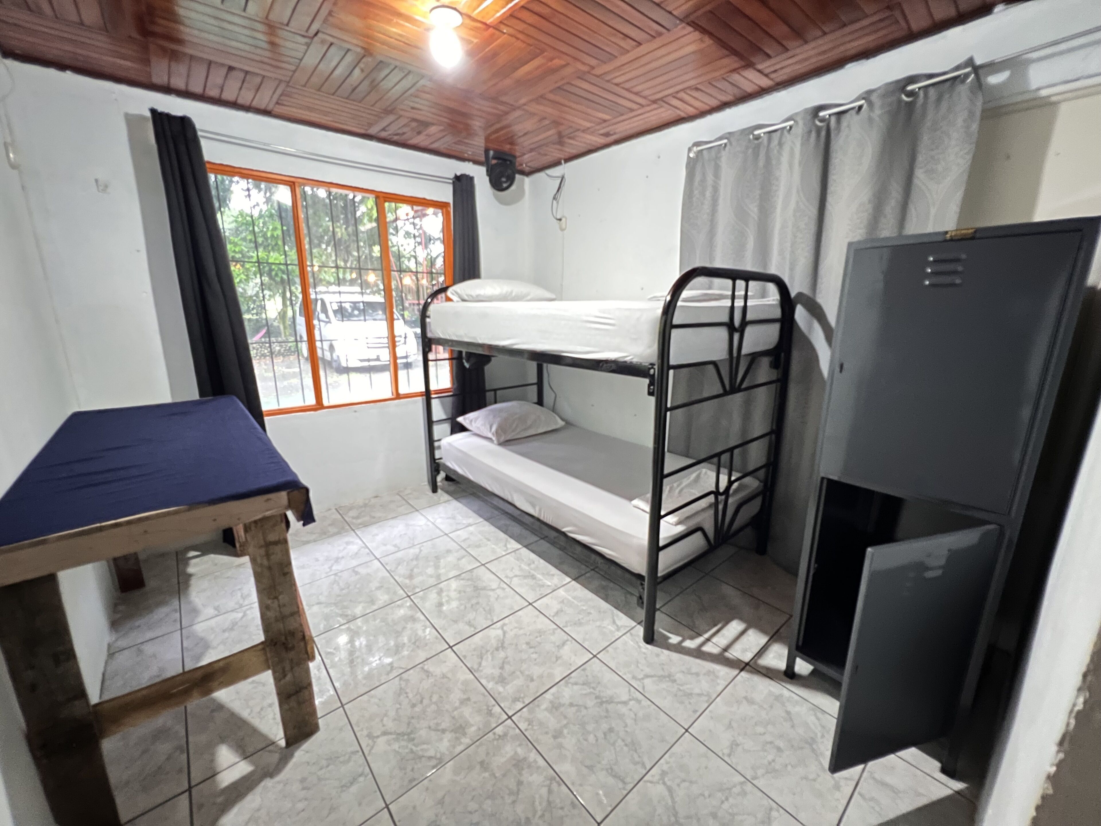 Un dormitorio con literas, una mesita pequeña, un refrigerador y una ventana con cortinas.