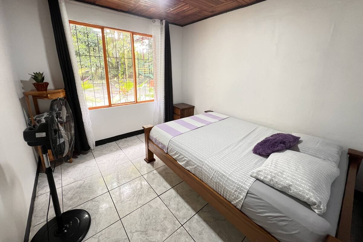 Une chambre à coucher avec un lit, un ventilateur sur pied, une petite table, une plante en pot et une fenêtre donnant sur un paysage verdoyant.