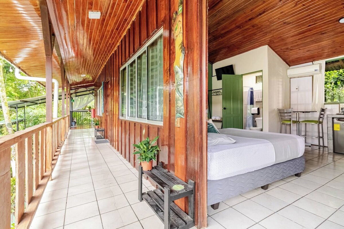 Uma cabana de madeira com varanda, cama, pequena cozinha e banheiro.
