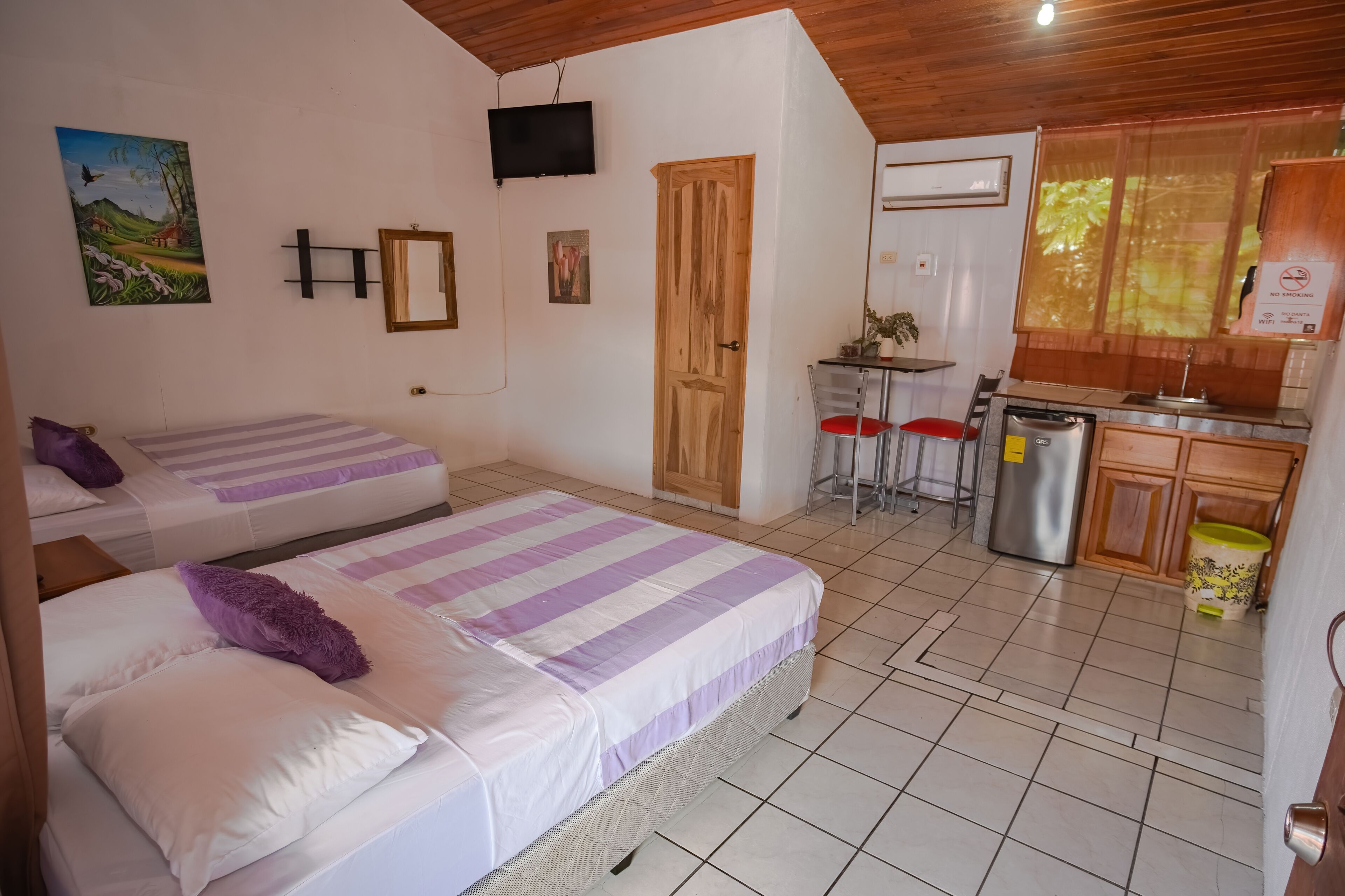 A room with two beds, a small kitchenette, a wooden ceiling, and a painting on the wall.
