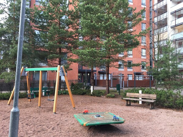 A playground with swings and a slide, surrounded by trees and a building in the background. - Nice apartment with sea view<br> (Helsinki)