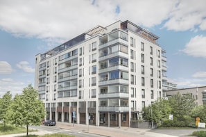 A multi-story residential building with balconies, a parking area, and a clear sky.