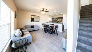 Amplia sala de estar con un sofá gris, mesa de comedor y sillas, una cocina al fondo y una escalera.