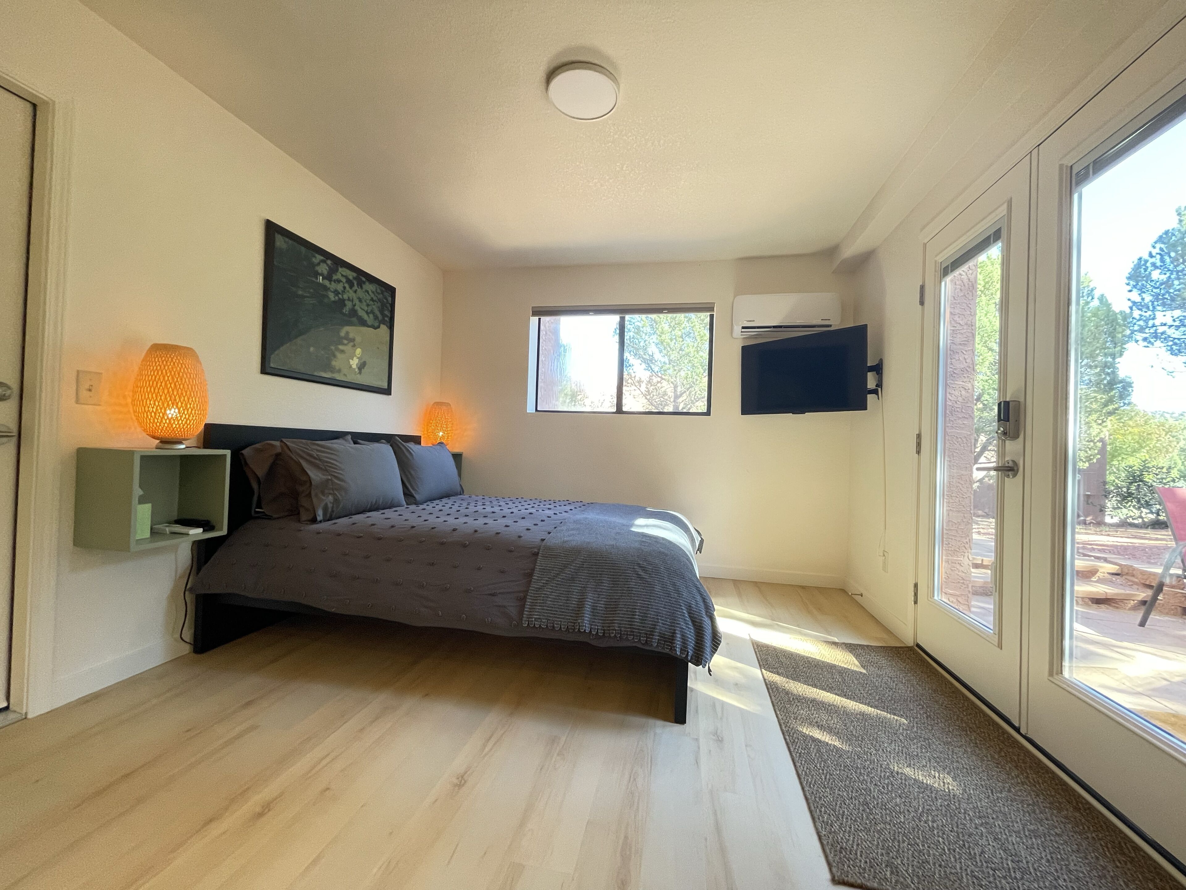 Une chambre à coucher comprenant un lit, des lampes de chevet, un téléviseur fixé au mur, une fenêtre et une porte donnant sur un espace extérieur.