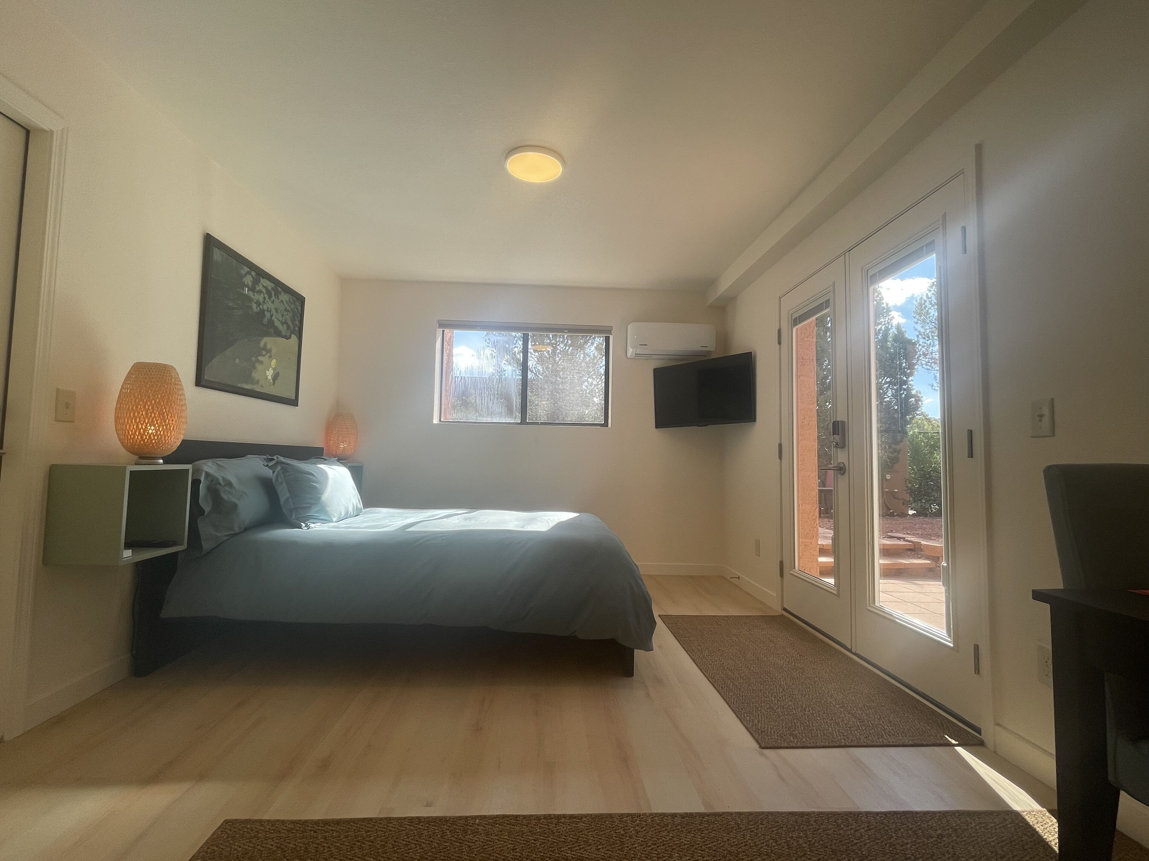 A bedroom with a bed, a bedside lamp, a flat-screen TV, and a window with a view of the outdoors.