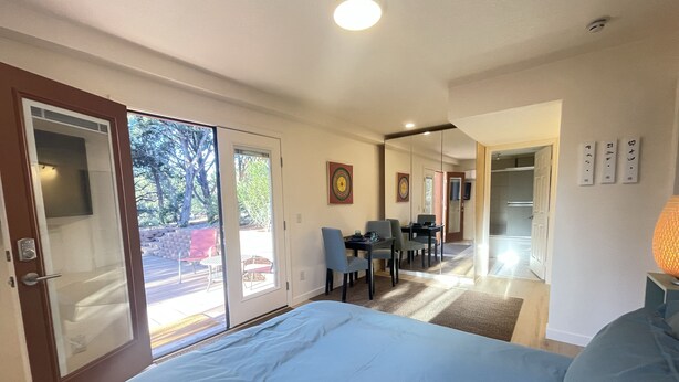 A modern hotel room with a large bed, a dining area with chairs, and a sliding glass door leading to an outdoor patio.
