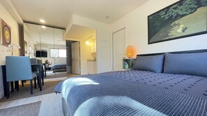 A modern bedroom with a large bed, a desk, and a chair. There is a door leading to another room, and a painting on the wall.