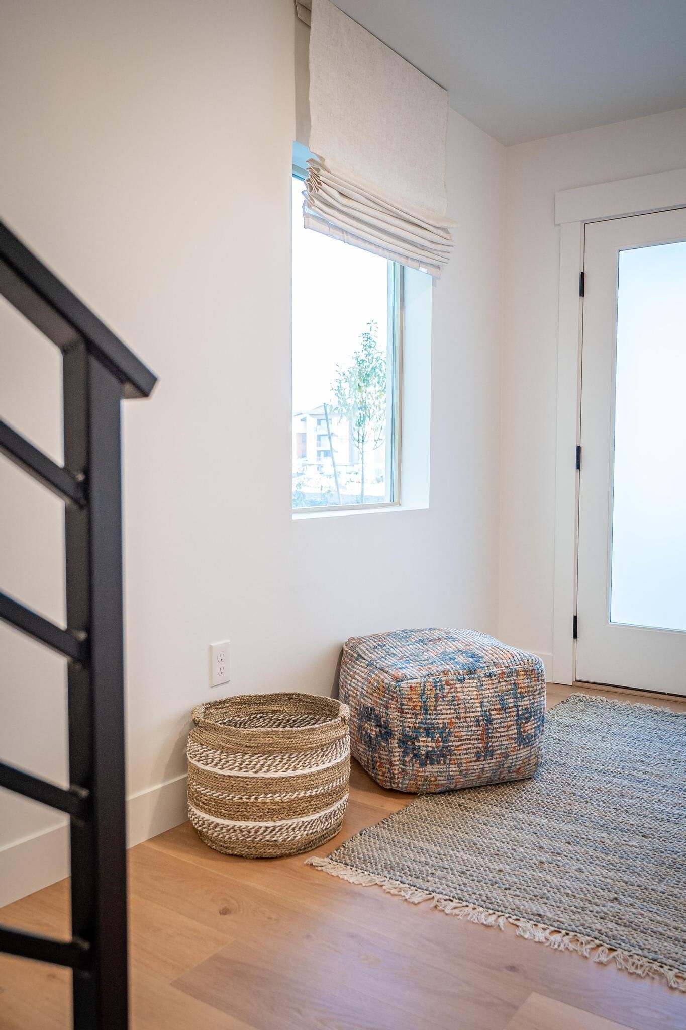 Ein Zimmer mit Holzboden, einem gewebten Korb, einem gemusterten Hocker, einem Fenster mit weißem Vorhang und einer Tür.