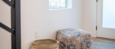 Ein Zimmer mit Holzboden, einem gewebten Korb, einem gemusterten Hocker, einem Fenster mit weißem Vorhang und einer Tür.