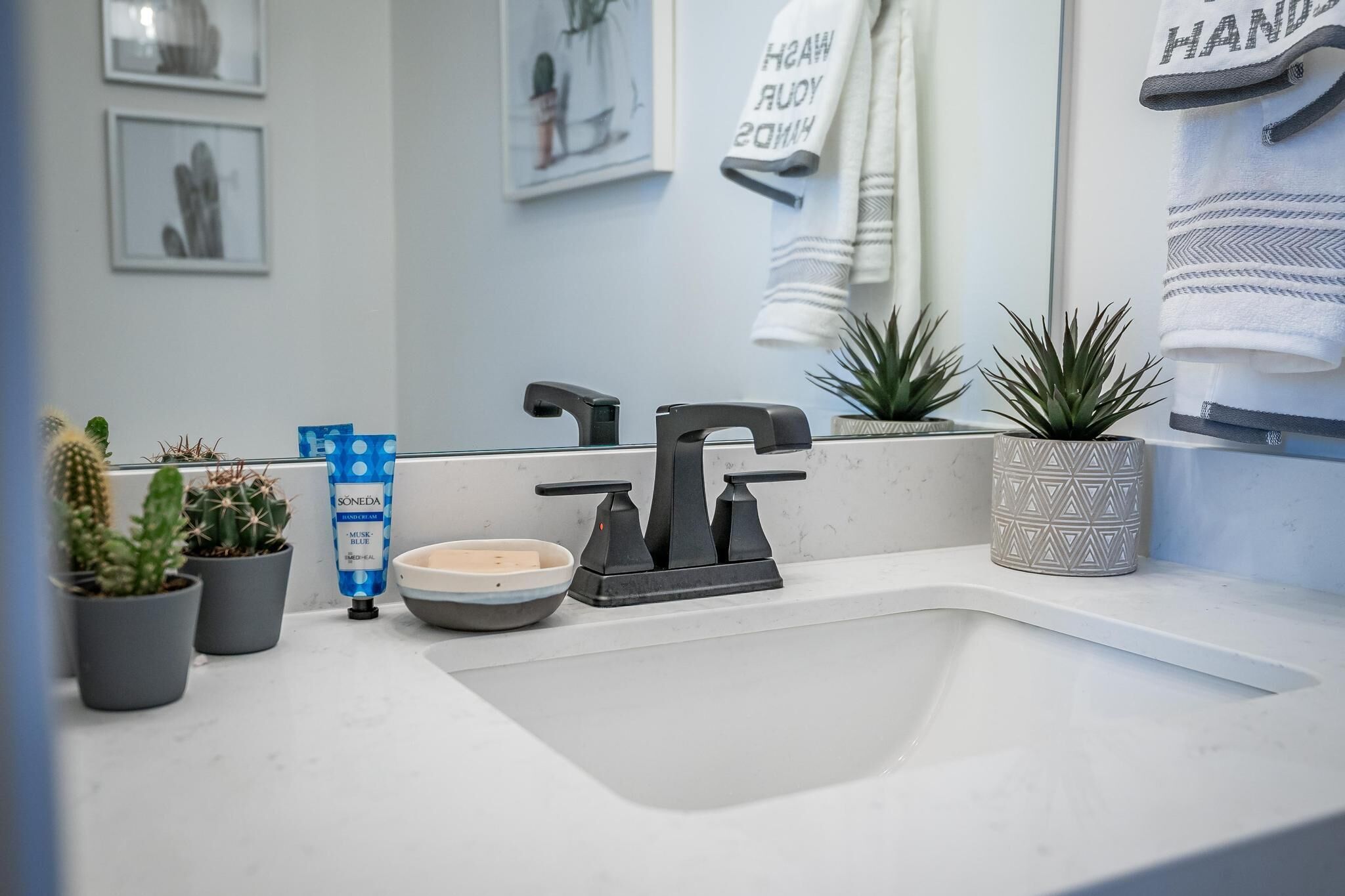 A modern bathroom with a white sink, black faucet, and a towel rack with a towel hanging on it. There are potted plants on the counter and a blue tube of lotion.