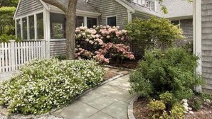 A residential house with a covered porch, a white picket fence, and a variety of flowering shrubs.