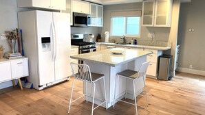 A modern kitchen with white cabinets, a central island with bar stools, stainless steel appliances, and a window providing natural light. - Treasured Bliss *  AC * Clean * Steps to Sand * Exclusive Patio (Newport Beach)