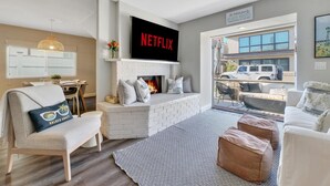 A living room with a fireplace, a TV mounted above it, and a view of the outside. - Treasured Bliss *  AC * Clean * Steps to Sand * Exclusive Patio (Newport Beach)