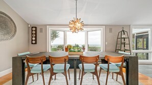 Un comedor con una mesa grande, cuatro sillas, una lámpara colgante y vistas al exterior a través de amplios ventanales.