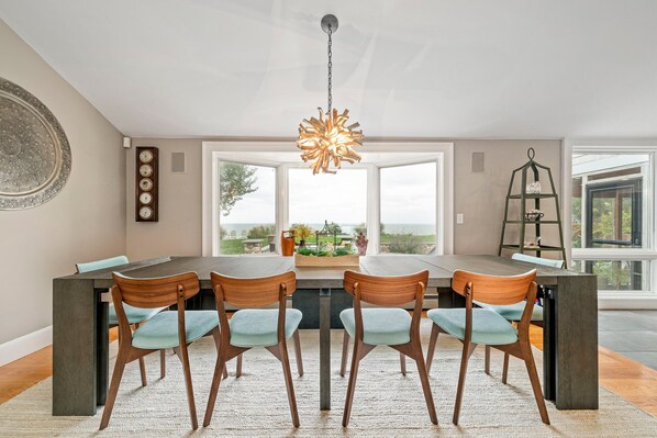 A dining area with a large table, four chairs, a hanging light fixture, and a view of the outdoors through large windows.