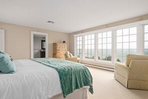 A bedroom with a large bed, a dresser, a chair, and a view of the ocean through large windows. - Stunning Oceanview: Walk to Private Beach (Plymouth)