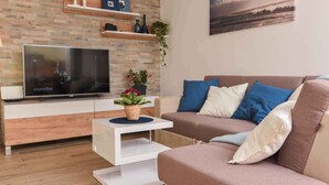 Ein modernes Wohnzimmer mit einem Flachbildfernseher, einem weißen Couchtisch, einem beige farbenen Sofa mit blauen und weißen Kissen sowie einer Wand mit Ziegelmuster und Regalen darüber.