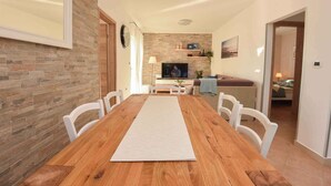 Ein Essbereich mit einem Holztisch, weißen Stühlen, einer Ziegelwand, einem Fernseher und einer Couch im Hintergrund.