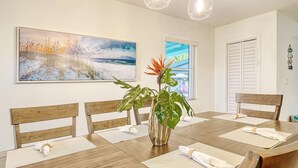 Uma área de refeições com mesa de madeira, cadeiras, uma pintura grande e uma planta.