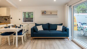Ein modernes Wohnzimmer mit einem dunkelblauen Sofa, weißen Stühlen, einem Esstisch mit blauen Schalen, einem kleinen Regal mit Büchern und einem Seestern sowie einem großen Fenster mit Blick auf Grünflächen.