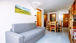 A modern living room with a grey sofa, a dining area with wooden chairs, and a wooden door leading to another room. - Cosy apartment for 5 guests with WIFI, hot tub and TV (Canazei)