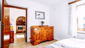 Un dormitori amb un taulell de fusta, un llit amb roba de llit estampada, un mirall a la paret i vista d'una cuina a través d'una porta oberta.