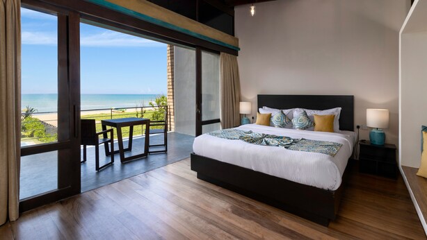 Une chambre avec un grand lit, un bureau et une chaise, une vue sur la plage et un ventilateur de plafond.