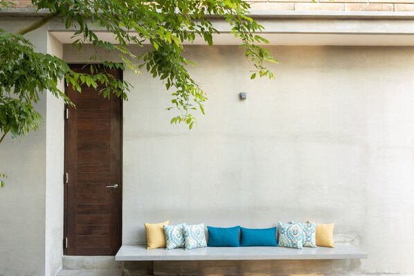 A bench with cushions against a plain wall, a door to the left, and a tree branch above.