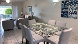 Een moderne woonkamer met een eettafel met glazen blad, omringd door zes grijze stoelen, een bank en een salontafel. Twee wandlampen en een inlijsting foto hangen aan de muur.