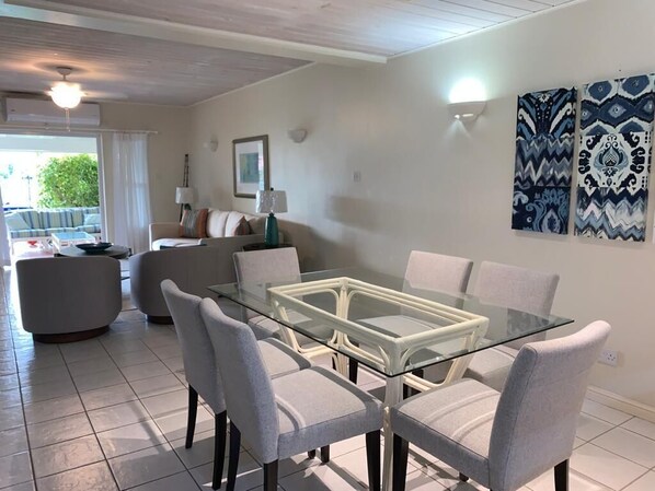 A modern living room with a glass-top dining table surrounded by six grey chairs, a sofa, and a coffee table. There are two wall-mounted lamps and a framed picture on the wall.