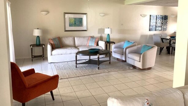 A living room with a white sofa, a teal armchair, and a red chair. There are two side tables with lamps, a ceiling fan, and a framed picture on the wall. - Bay View #7 is a 1-bed, 1-bath waterfront townhouse in a gated community. (Rodney Bay)
