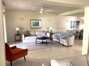 A living room with a white sofa, a teal armchair, and a red chair. There are two side tables with lamps, a ceiling fan, and a framed picture on the wall.