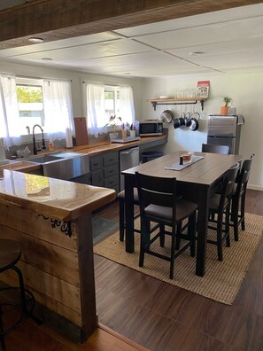 Een moderne keuken met een centraal eiland, een houten eettafel en stoelen. Er is een magnetron, een koelkast en een plank met keukengerei.