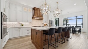 Una cocina moderna con armarios blancos, una isla de madera, taburetes negros y ventanas grandes con vistas.