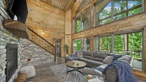 A spacious living area with a stone fireplace, a leather sofa, and a large window overlooking a forest.