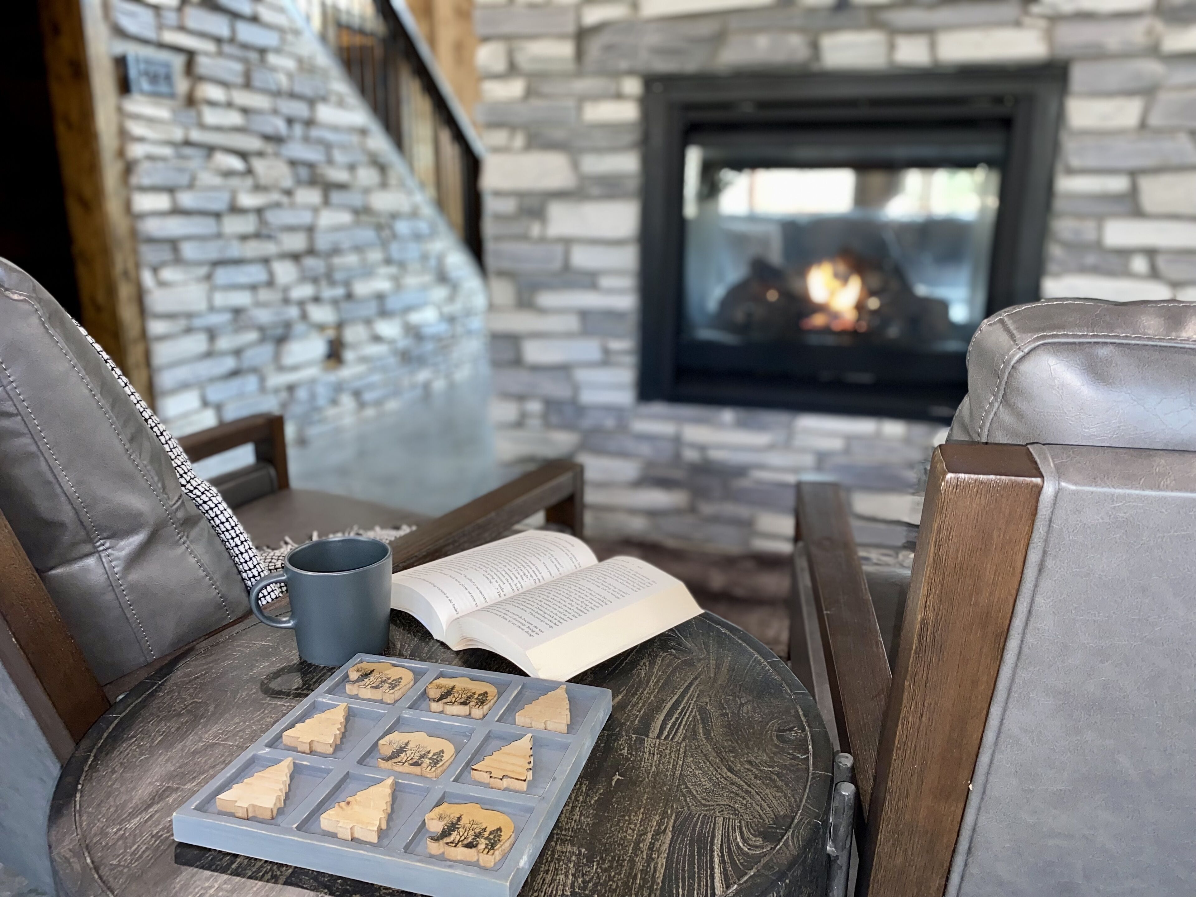 A cozy seating area with leather chairs, a wooden table, a book, a mug, and a tray of wooden Christmas trees. A stone wall and a fireplace with a fire are in the background.