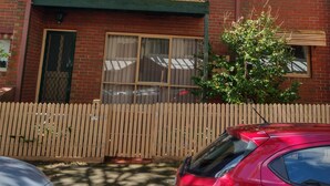 Un edificio in mattoni di due piani con un balcone, una recinzione di paletti e un'auto rossa parcheggiata davanti.