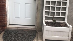 Una puerta blanca con un ventanal pequeño, una cámara de seguridad encima y una enrejada blanca con una maceta junto a la puerta.
