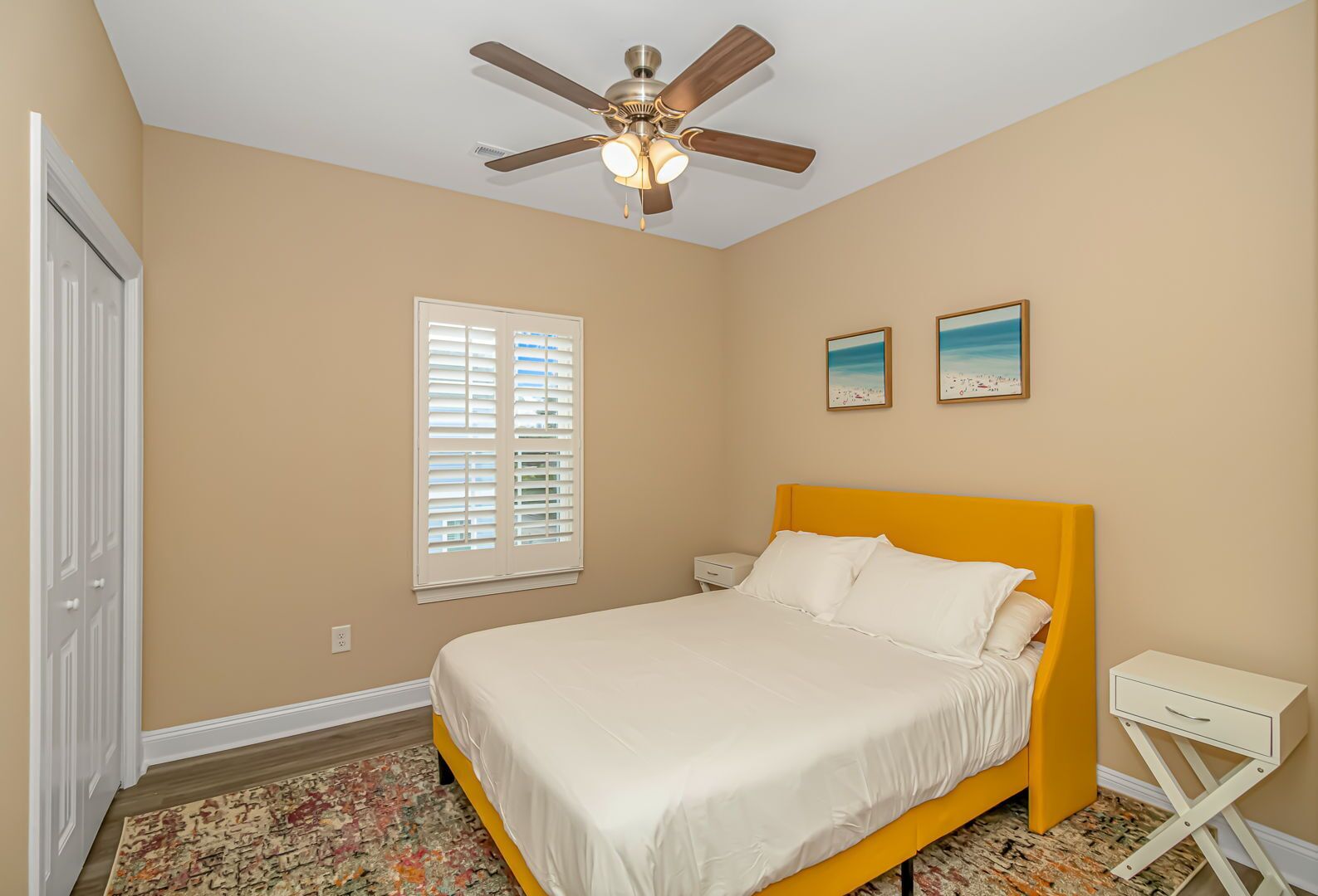 A bedroom with a bed, a ceiling fan, a window with blinds, and two framed pictures on the wall.