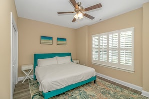 Quarto com cama verde-água, roupa de cama branca, ventilador de teto, duas obras de arte nas paredes, janela com persianas e tapete estampado.