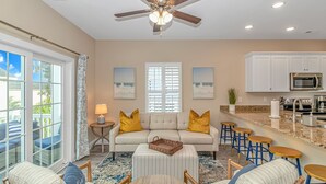 A spacious living room with a sofa, ottoman, and bar stools, a kitchen with white cabinets and appliances, and a ceiling fan.