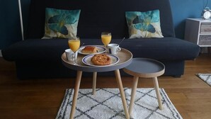 Un salon chaleureux avec un canapé bleu foncé, deux coussins à motifs floraux, une petite table ronde sur laquelle sont disposés des éléments pour le petit-déjeuner, et une table d’appoint avec une lampe.