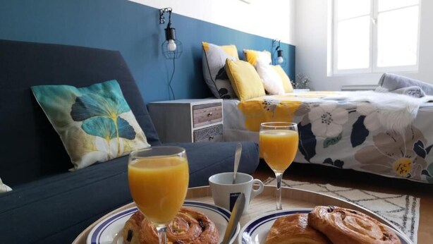 Un plateau de petit-déjeuner avec des croissants et du jus d’orange, un lit recouvert d’une couette à motifs floraux et un mur bleu orné d’un coussin en forme de fleur.