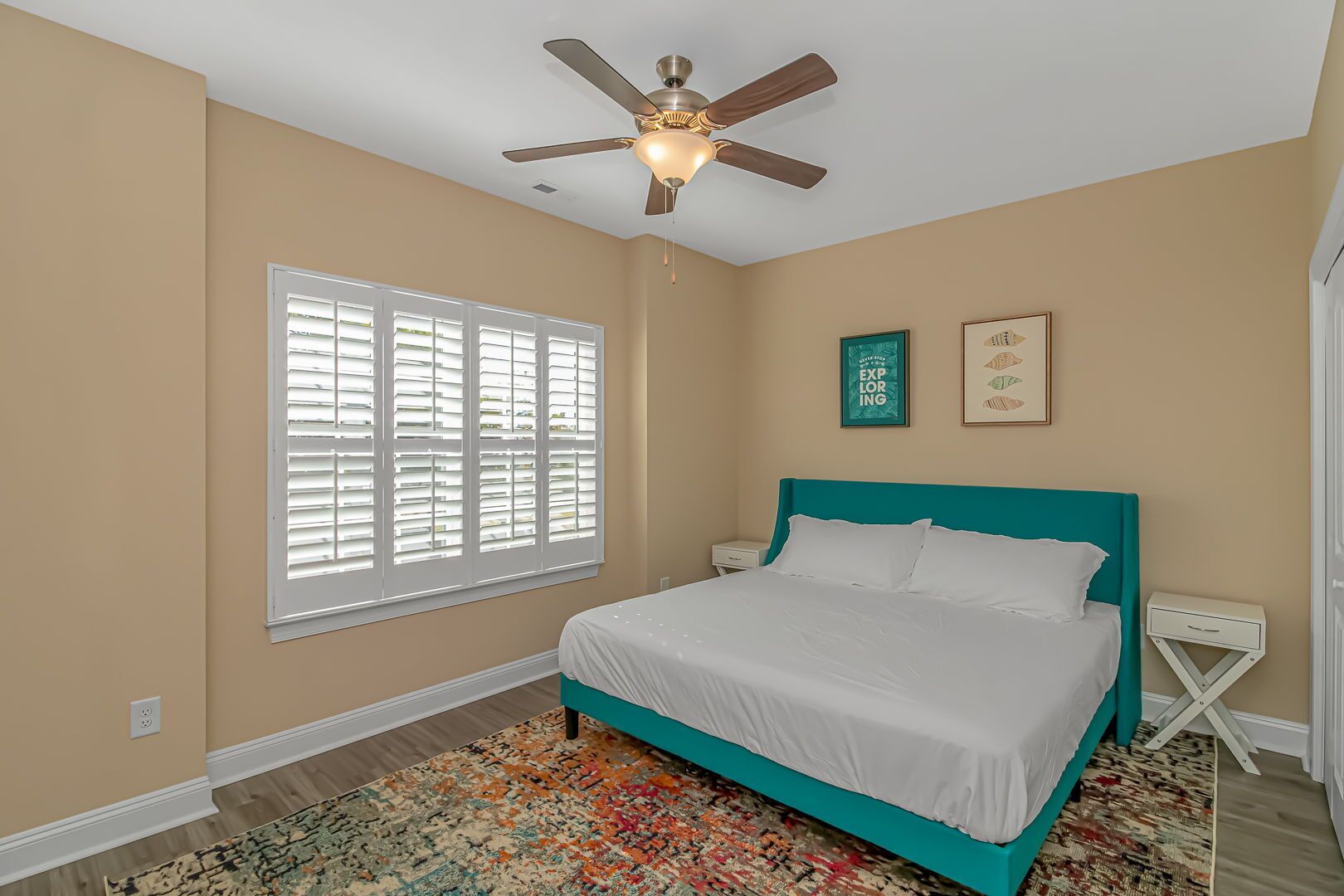 Un dormitorio con cabecera color turquesa, ropa de cama blanca, ventilador de techo, ventana con persianas y alfombra estampada.