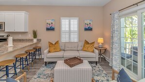 A living room with a sofa, two side tables, a coffee table, and a kitchen area with a countertop and bar stools. - PRIVATE HOT TUB, steps from the beach, boardwalk and many other attractions. (Myrtle Beach)